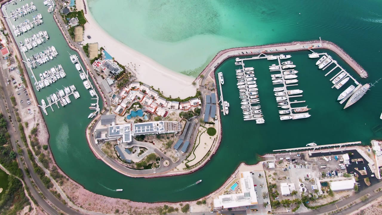 Aerial marina view with white boats, sandy beach, buildings, road, and small boat wake in the channel.