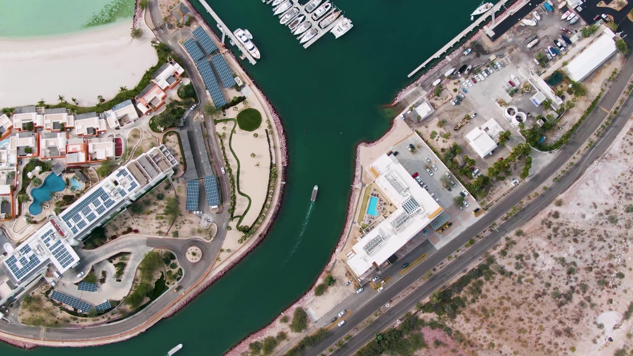Aerial overhead frame in La Paz with the boat farther along the green canal, docked marina boats at the top, solar-panel roofs, a road, sandy lots, and no visible people.