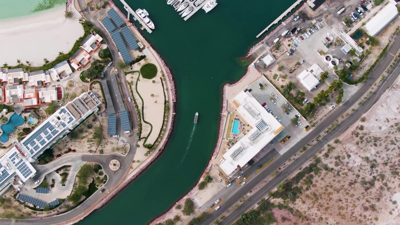 Top-down La Paz marina canal frame with the boat and wake centered in the waterway, white structures, docked boats, roadway traffic, sandy terrain, and no visible people.
