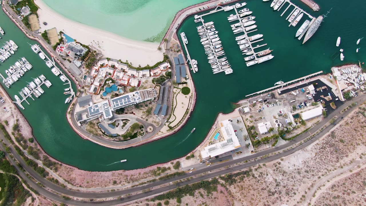 Top-down aerial frame of a La Paz marina with rows of docked boats, a pale sandy beach, curved seawall, road, low buildings, and no visible people.
