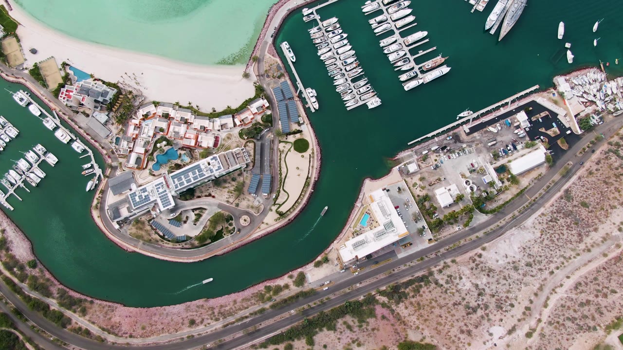 La Paz aerial frame showing a small boat entering the lower channel, marina docks packed with white vessels, a bright beach curve, nearby buildings, and no visible people.