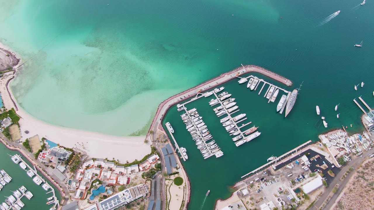 High overhead aerial frame of a La Paz marina beside a curved sandy beach, with docked boats in teal water, pale turquoise shallows above, coastal buildings below, and no visible people.