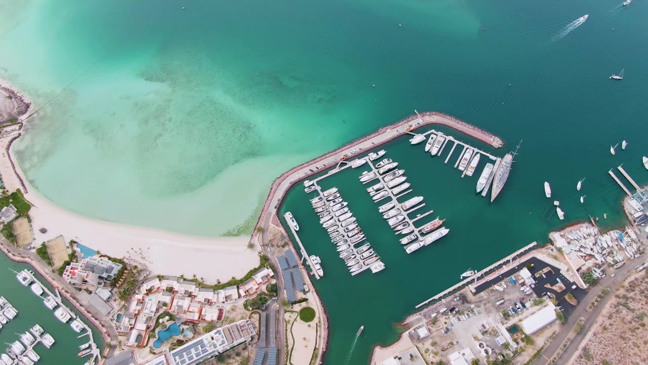 High aerial view of the La Paz shoreline with the marina and breakwater angled through the center, beach and buildings along the lower edge, scattered small boats offshore, and no visible people.