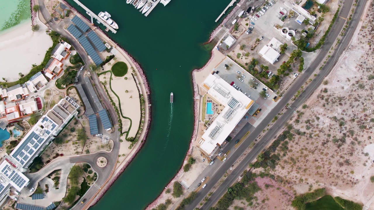Overhead view in La Paz, Baja California Sur showing a green marina channel, pale beach, docked boats, waterfront buildings, road, scrubby desert ground, and no visible people.