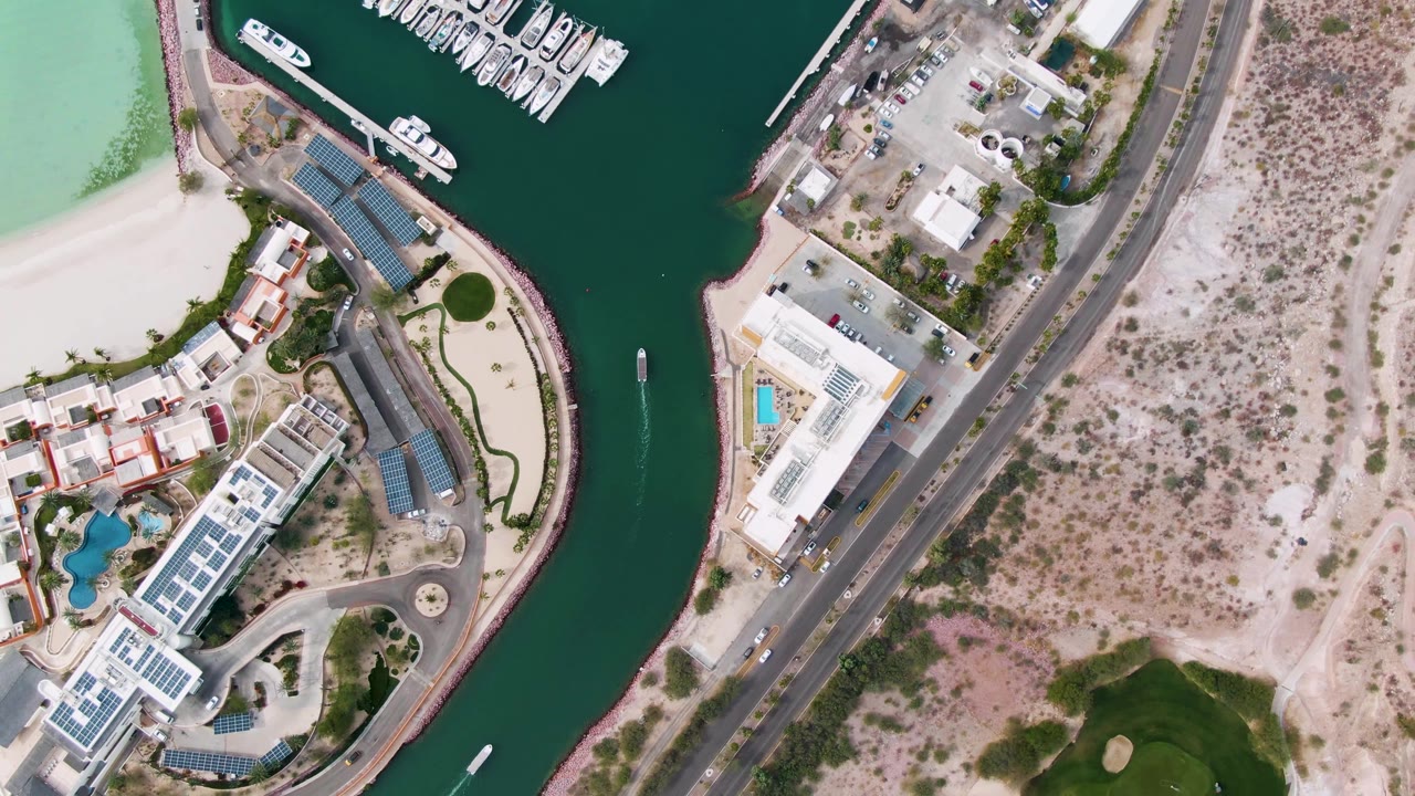 Top-down view in La Paz, Baja California Sur showing turquoise beach water, a green marina channel, docked white boats, a road through scrubland, buildings, and no visible people.
