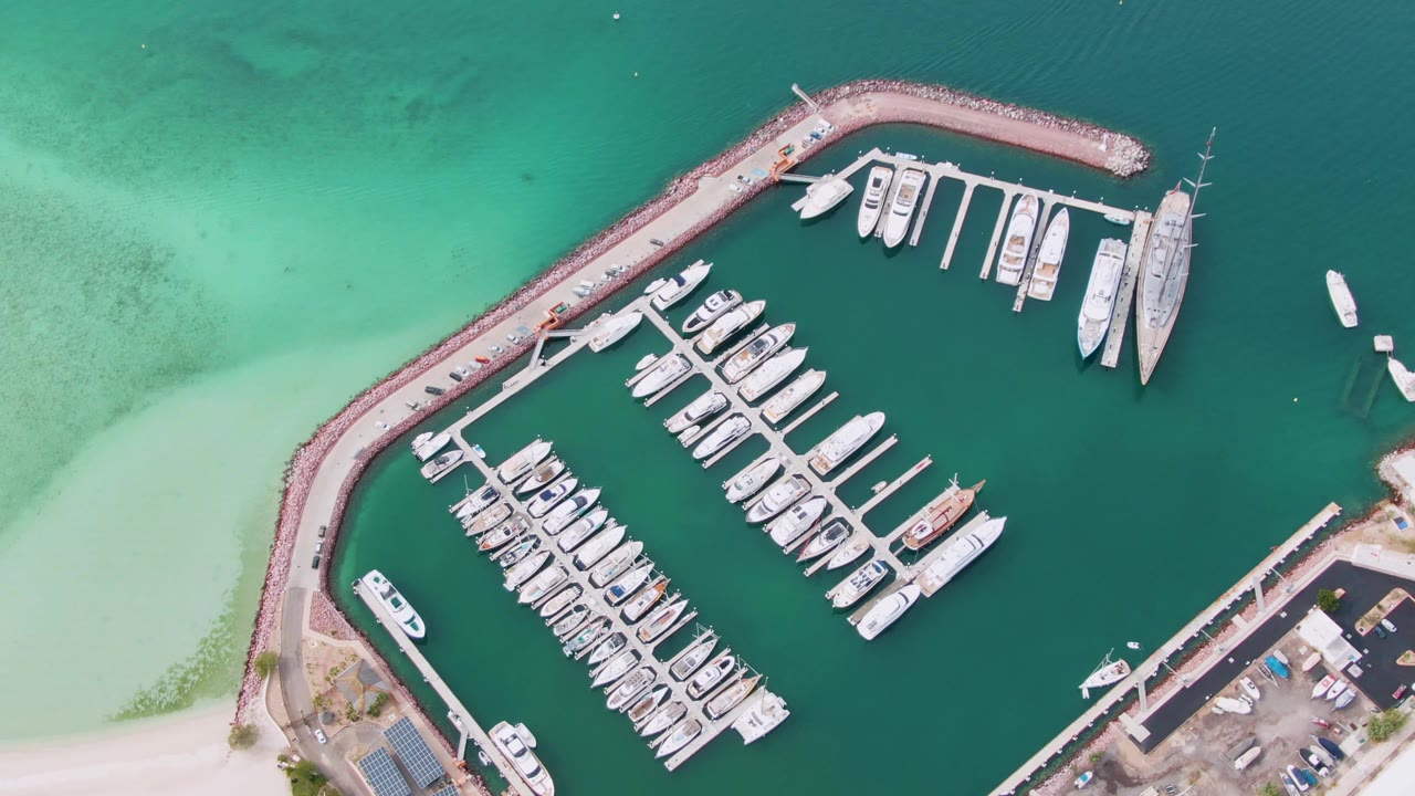 Top-down aerial view of a La Paz marina with white boats in slips, a curved rock breakwater, pale turquoise shallows at left, deeper green harbor water, and no visible people.