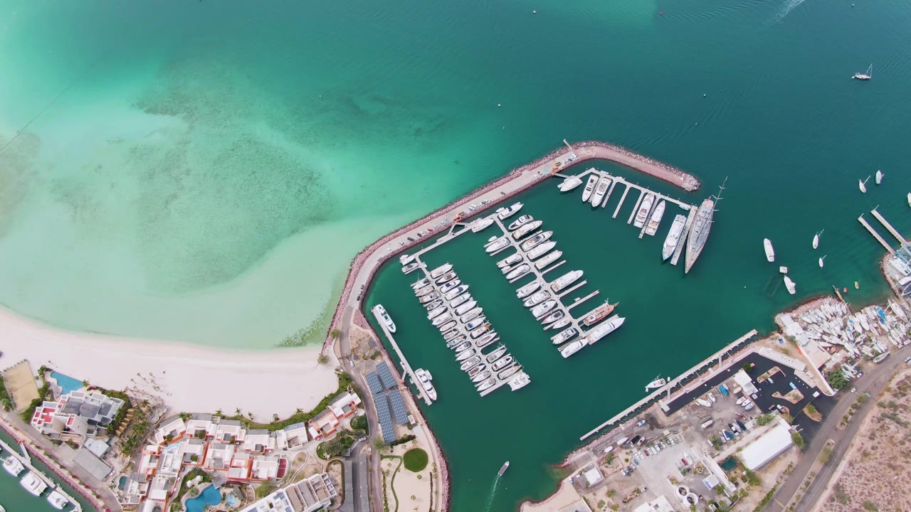 Aerial overhead frame of a La Paz marina with rows of docked yachts inside a curved breakwater, pale beach at left, turquoise water above, and no visible people.