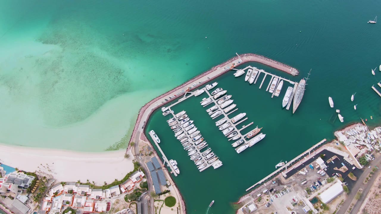 Curved marina breakwater with yachts, beach, and clear green-blue water.