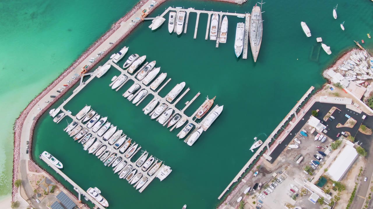 Overhead La Paz marina view with docked white boats in green water, turquoise shallows outside the breakwater, shore parking to the right, and no visible people.