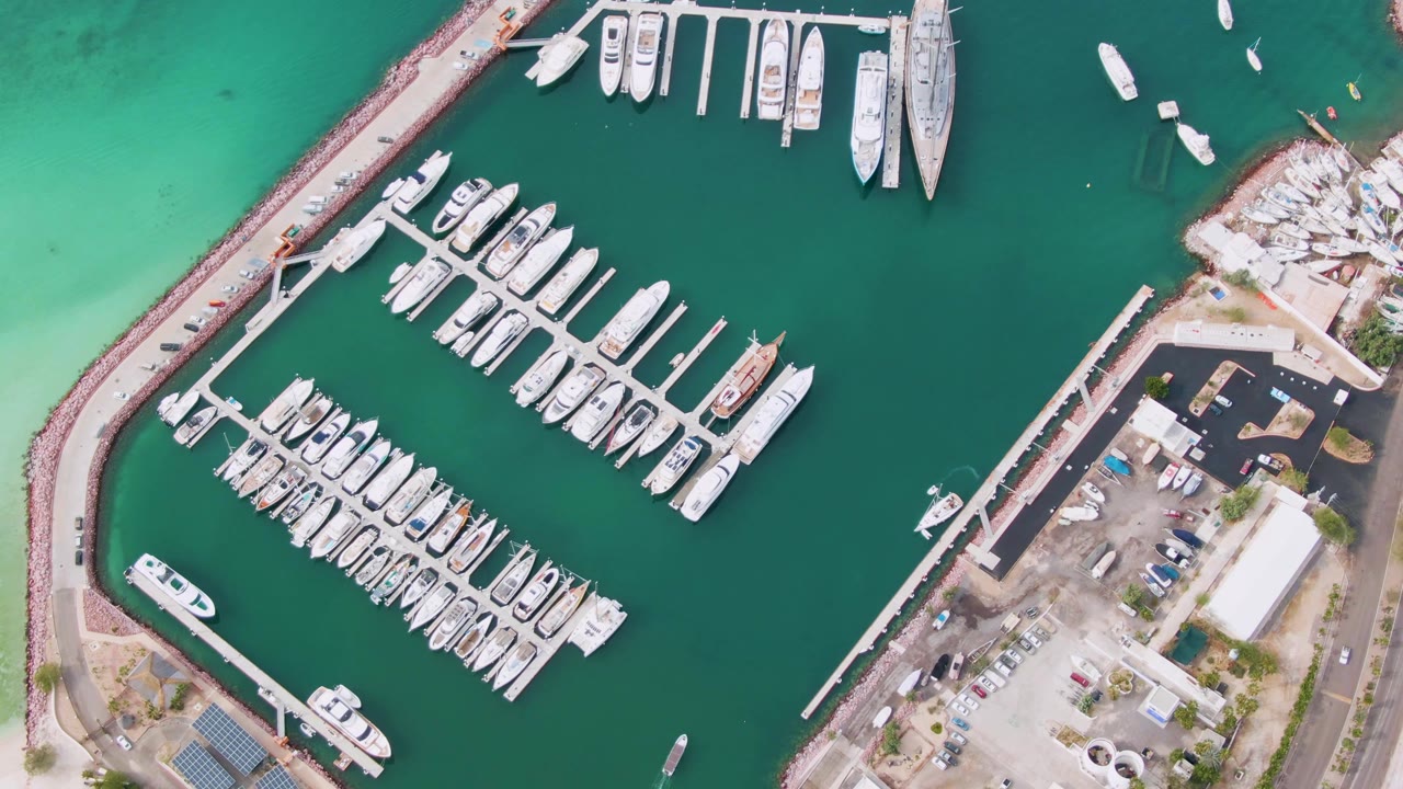 Top-down drone frame over the La Paz harbor marina, showing parallel docks packed with boats, a long breakwater, a small vessel in the water, shore-side cars, and no visible people.