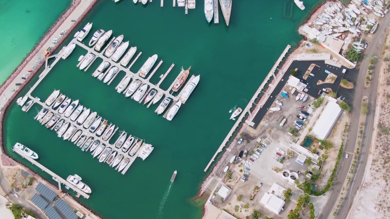 Overhead view of a La Paz marina with rows of docked white boats, green water, shoreline pavement, a nearby road, and no visible people at the start of the clip.