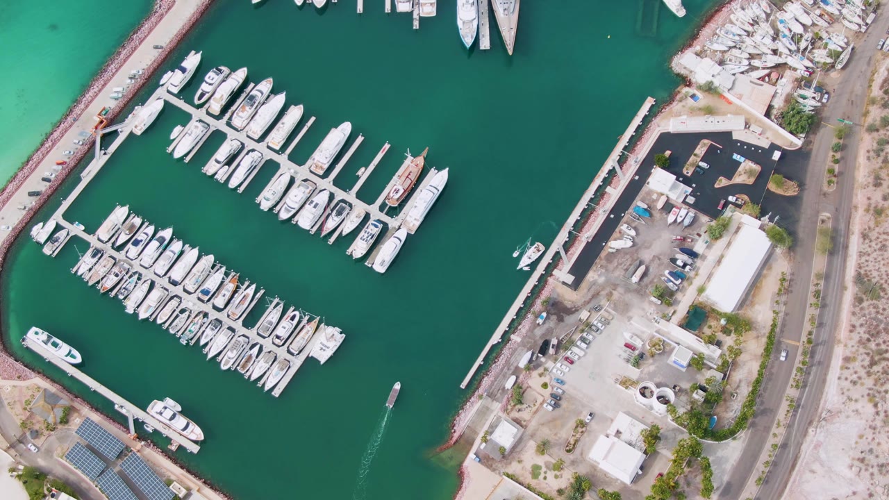Aerial frame over the La Paz marina showing packed dock rows, a small boat wake in green water, a parking lot, low buildings, road edge, and no visible people.