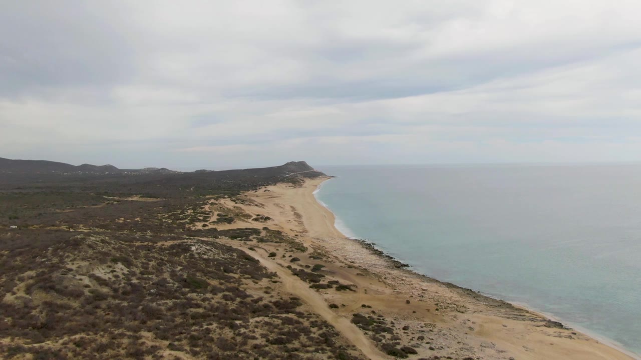Punta Gorda Beach aerial frame with inland scrubland spread across the foreground, a sandy beach tracing the waterline, distant low hills, muted overcast light, and no visible people.