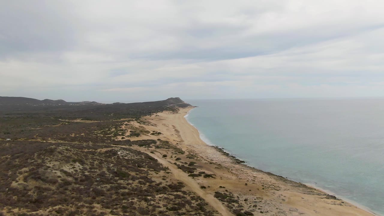 Mid-clip aerial view of Punta Gorda Beach showing broad scrub-covered dunes in the foreground, a tan beach narrowing toward the distance, calm ocean water, and no visible people.