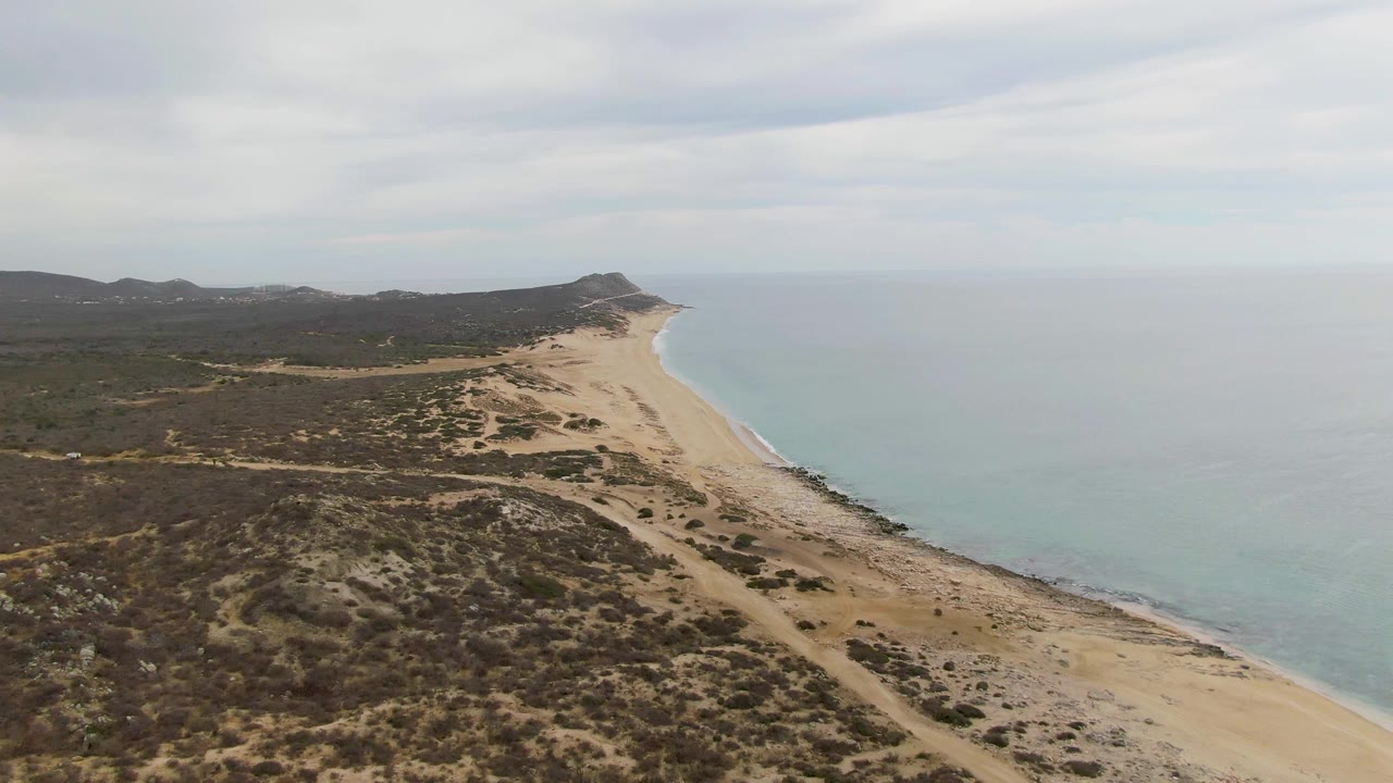 Punta Gorda Beach aerial frame showing the beach stretching toward a low headland, with scrub-covered dunes in the foreground and no visible people.