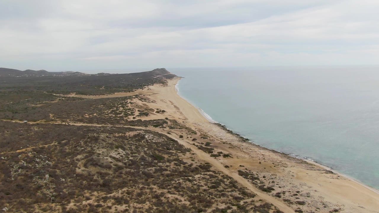 High coastal view with sandy beach, calm sea, scrubland, and distant hills
