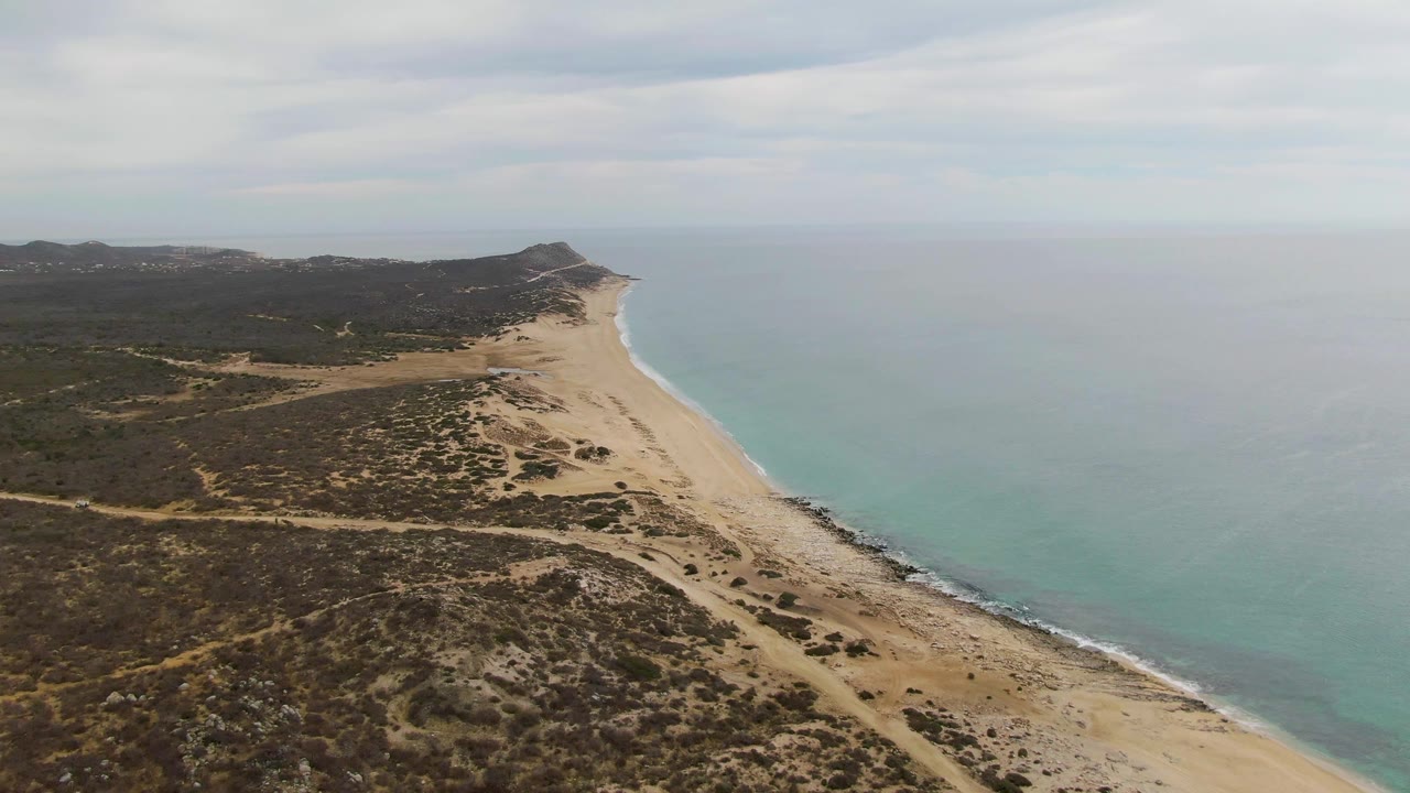 Aerial view of sandy beach, turquoise water, scrubland, and distant low hills.