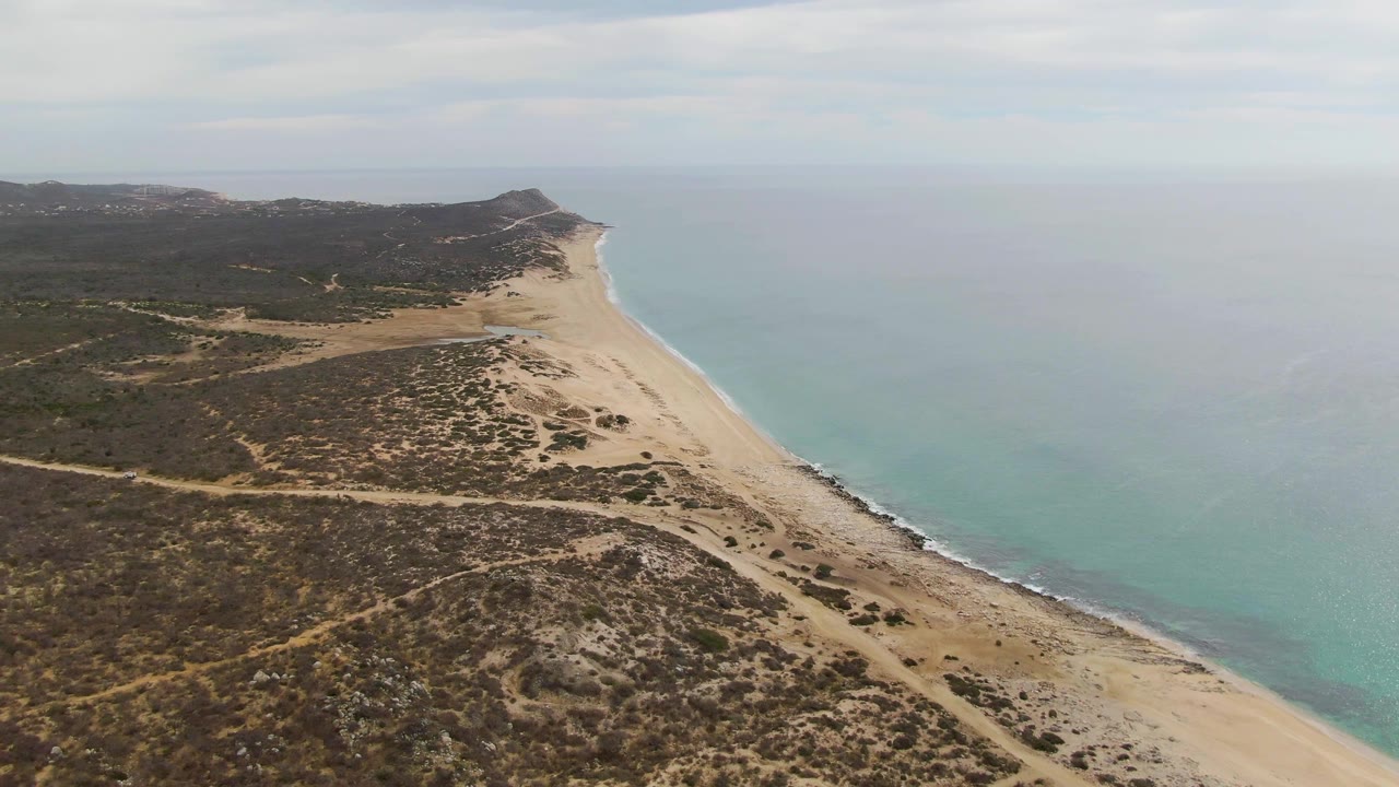 Aerial frame over Punta Gorda Beach, Baja California Sur, with pale sand curving beside turquoise water, dry shrubland in the foreground, and no visible people.