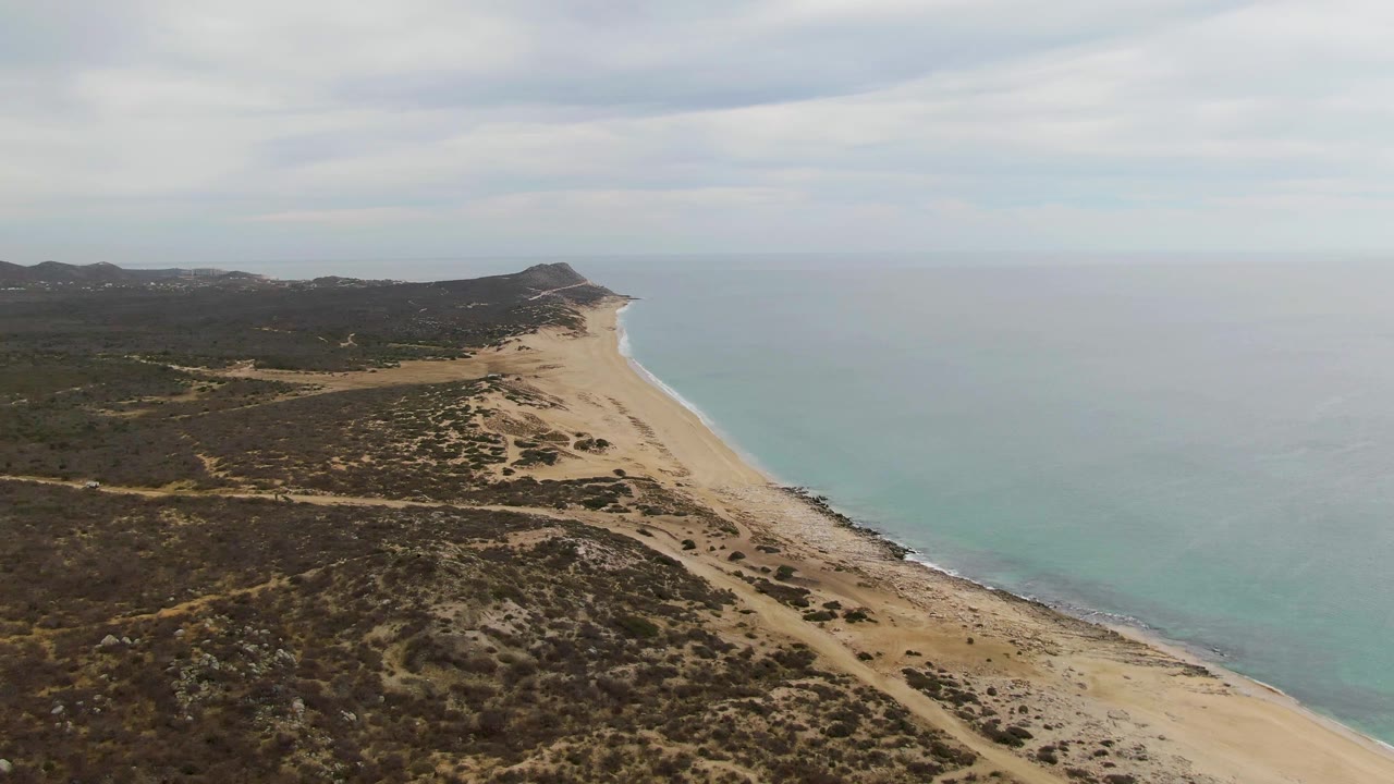 Aerial view at Punta Gorda Beach showing a long sandy strand between dark low vegetation and lighter turquoise water, with the coastline narrowing toward a headland and no visible people.