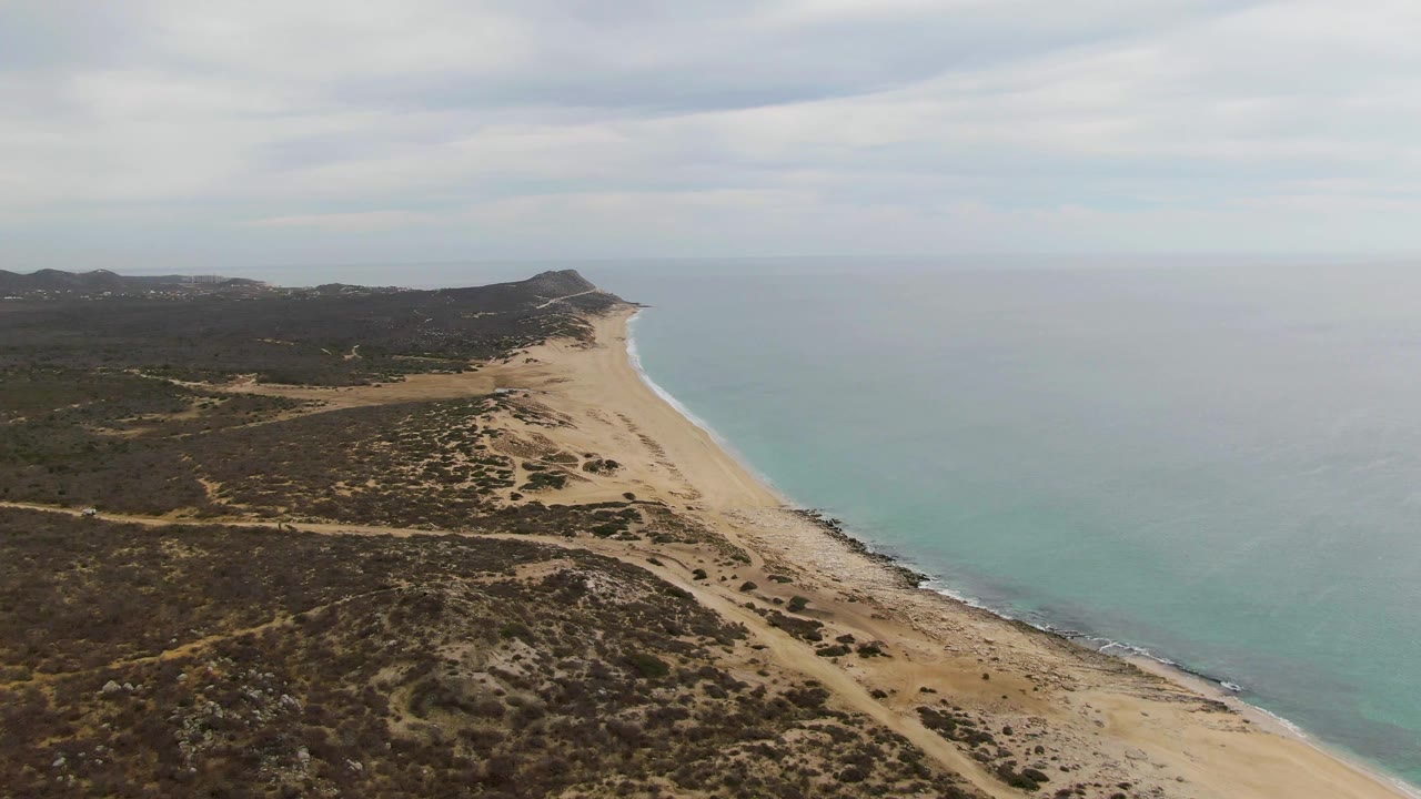 Aerial frame from Punta Gorda Beach with broad scrubland in the foreground, an empty sandy shoreline along the right edge, blue-green ocean water, distant hills, and no visible people.