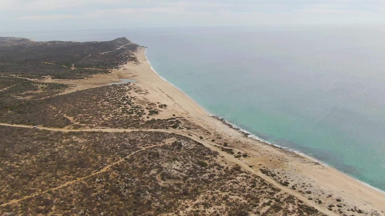 Aerial view over Punta Gorda Beach in Baja California Sur with scrubby dunes, pale sand, calm blue-green water, and no visible people at the start of the clip.