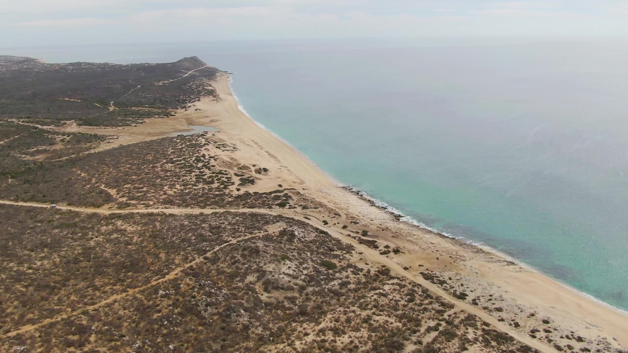 Aerial frame of Punta Gorda Beach showing the long sand strip, inland dune vegetation, faint dirt tracks, calm ocean, and no visible people one second into the clip.