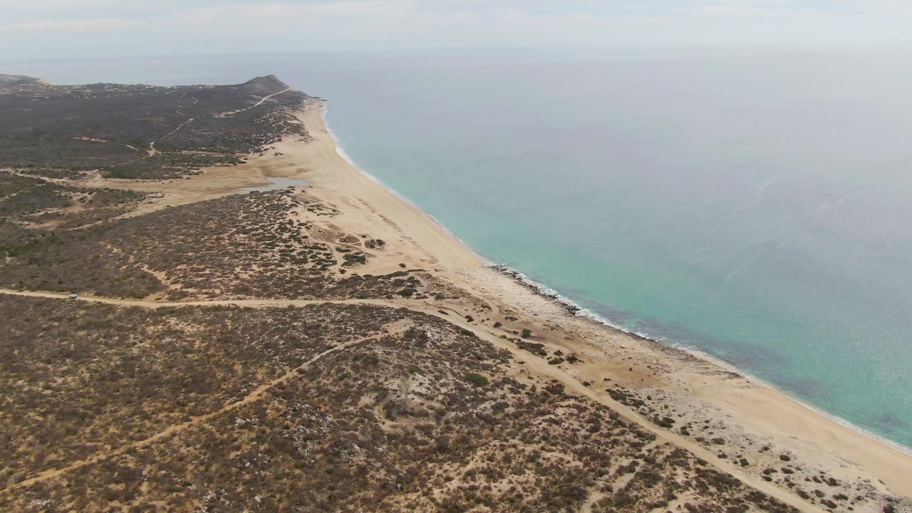 Oblique aerial view along Punta Gorda Beach with the sandy shore angling toward a distant coastal point, muted turquoise water on the right, and no visible people.