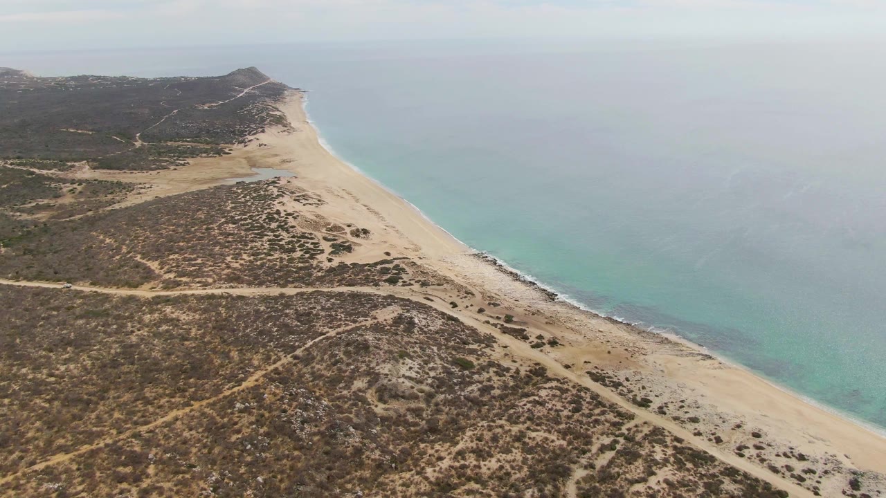 Wide aerial of beach, dune scrub, and calm ocean under hazy sky.