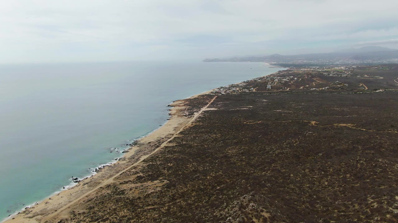 Wide aerial coastline with beach road, dry terrain, ocean, and hazy hills