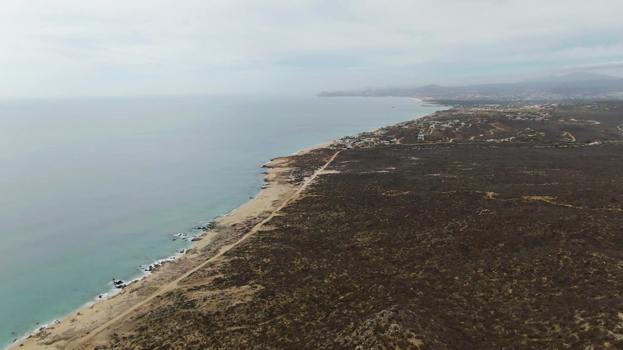 Mid-clip aerial frame at Punta Gorda Beach showing a long sandy shore, turquoise-gray water, a coastal road, dark scrub terrain, distant buildings near the hills, and no visible people.