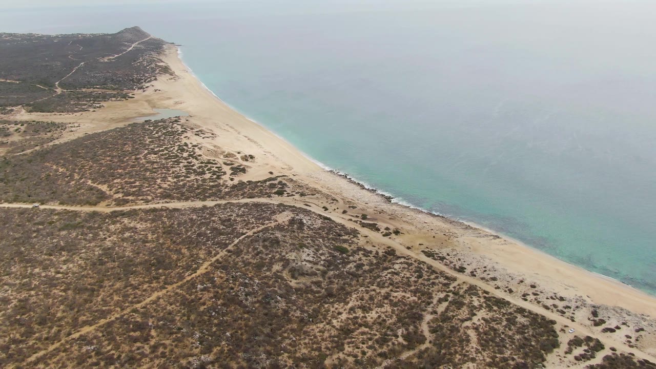 Wide aerial coastline with sandy beach, low surf, and brown-green dunes.