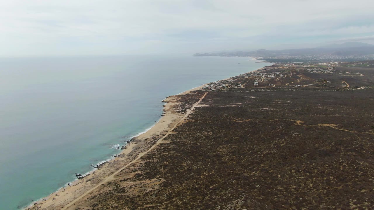 Punta Gorda Beach from above, with calm ocean on the left, a narrow sandy beach and dirt road below, dark coastal vegetation to the right, distant hills, and no visible people.