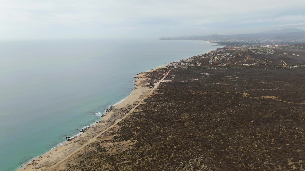 High coastal view with open water, curving shore, road, scrubland, and distant hills.