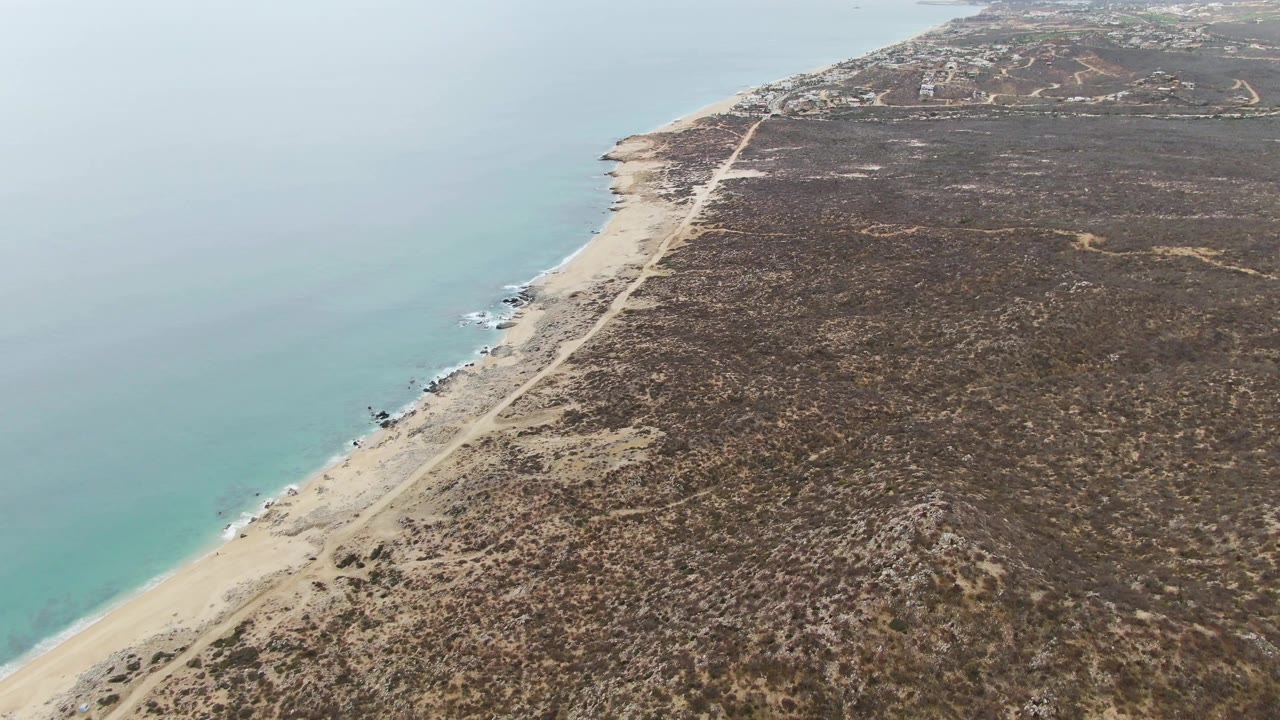 Aerial frame of a narrow beach and road beside pale blue ocean.