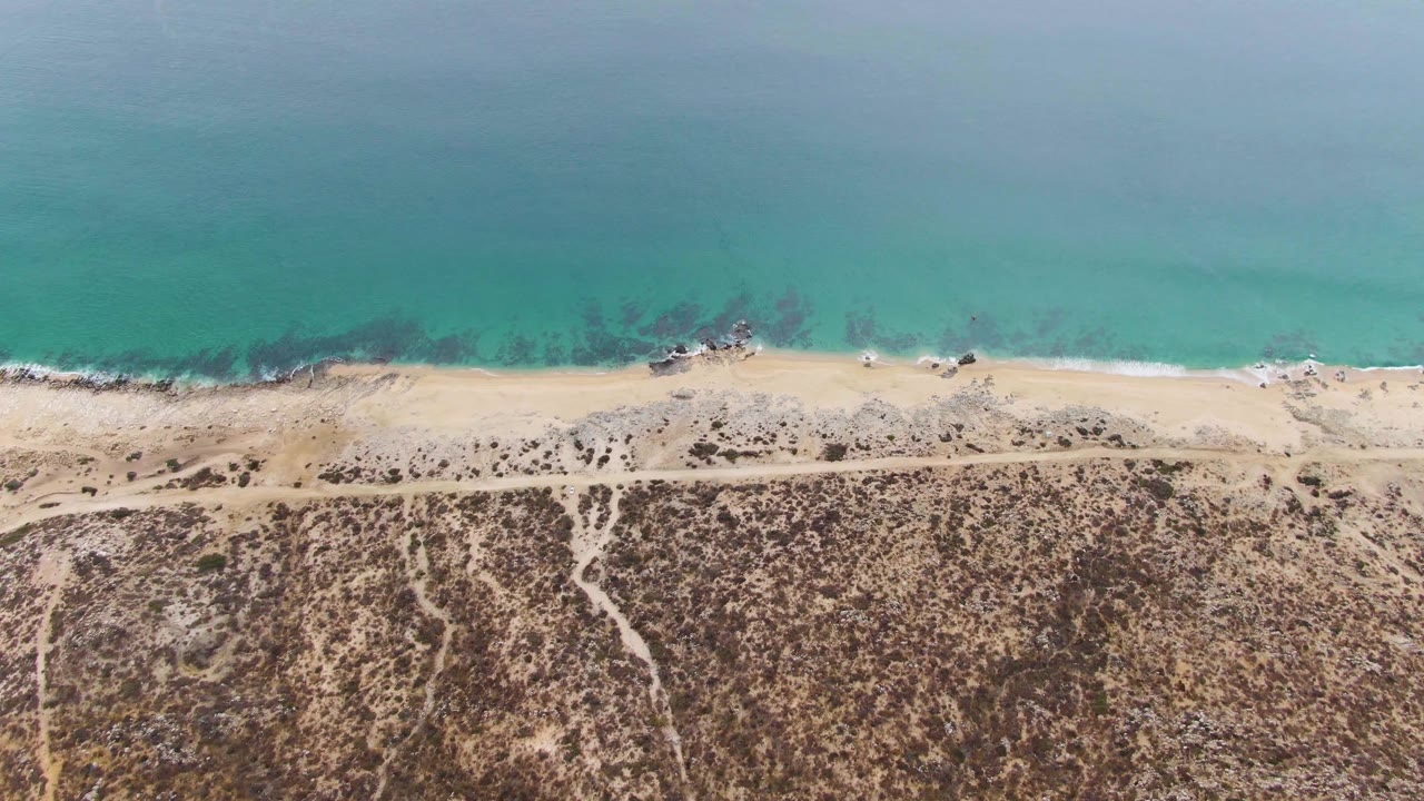 Aerial view of turquoise ocean, sandy beach, dirt road, and dry coastal terrain.
