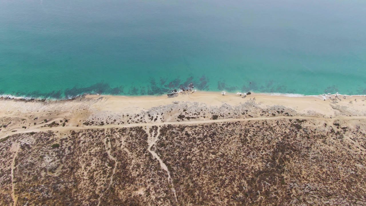 Aerial frame from Punta Gorda Beach showing a broad band of blue-green ocean, a narrow sandy shoreline, scattered dark rocks in the shallows, branching inland paths, and no visible people.