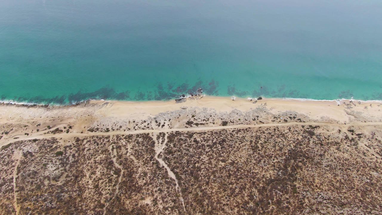 Mid-clip aerial view at Punta Gorda Beach with calm turquoise water, a thin white wave edge, pale sand, dark rocks near the shore, a straight dirt road, dry scrubland, and no visible people.
