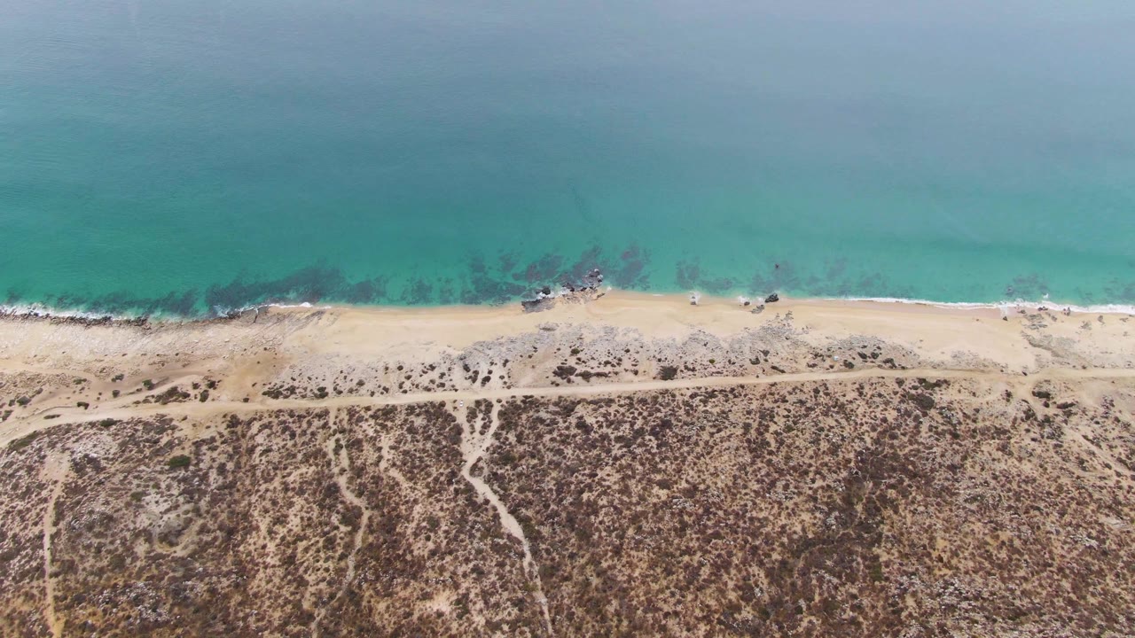 Aerial frame over Punta Gorda Beach with the turquoise ocean filling the top, small waves touching the sand, dark shallow-water rocks, a parallel dirt track, dry inland terrain, and no visible people.