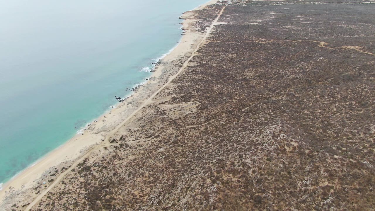 Drone-height frame of Punta Gorda Beach showing the road near the sand, faint surf against rocks, broad dry terrain inland, and no visible people.