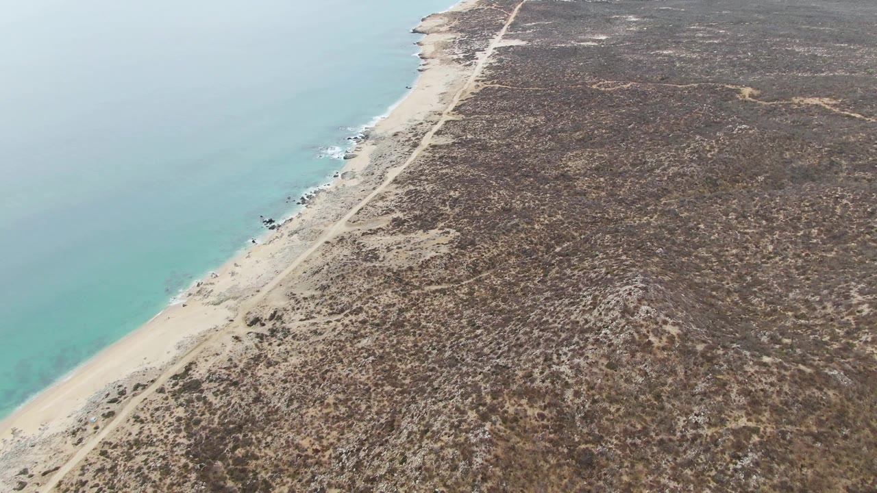 Mid-clip aerial frame at Punta Gorda Beach with turquoise water filling the left side, a narrow sand strip, the dirt road inland, dry brush, and no visible people.