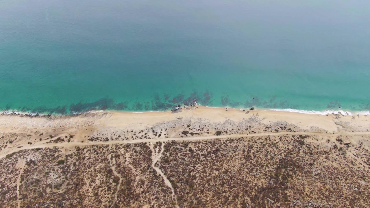 Overhead view of turquoise ocean, sandy beach, dirt road, and scrub dunes.