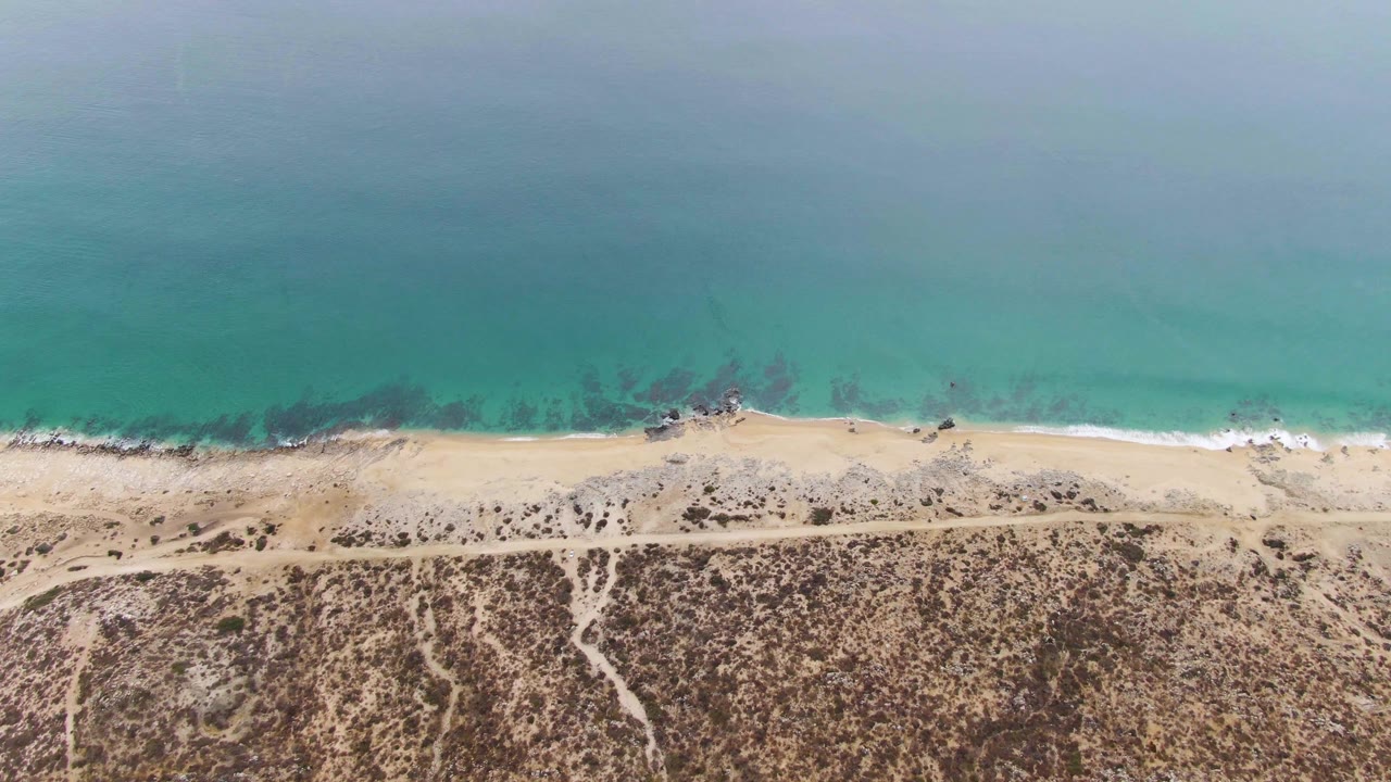 Aerial view over Punta Gorda Beach with the beach road running parallel to the shore, turquoise ocean water above the surf line, dry vegetation below, and no visible people.