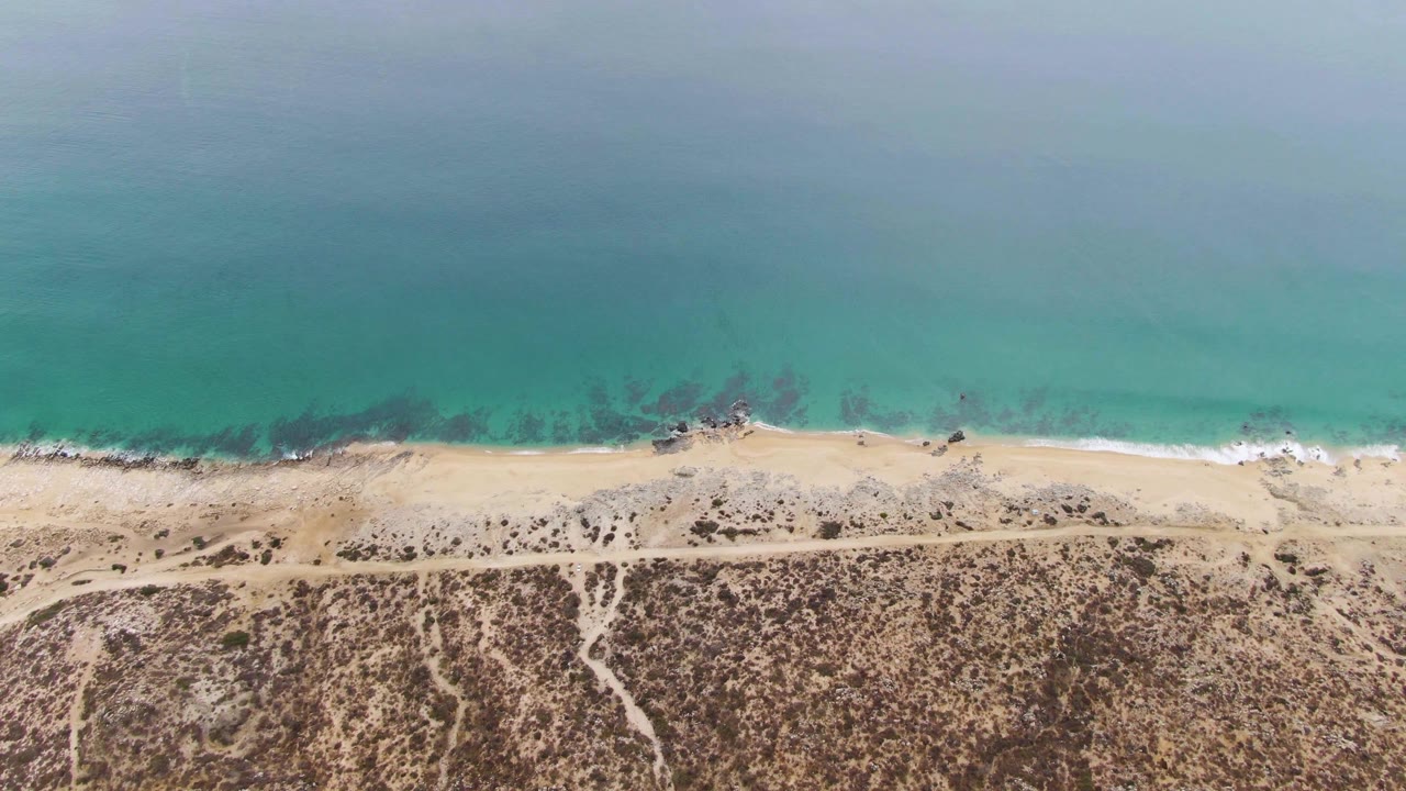 Top-down frame at Punta Gorda Beach showing blue-green ocean, a narrow sandy shore, scattered dark rocks near the waterline, dirt tracks through scrub dunes, and no visible people.