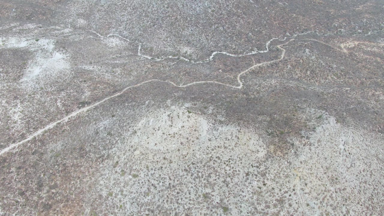 Aerial view over Punta Gorda Beach, Baja California Sur, with the winding dirt road near the upper portion of the frame and light sandy desert ground below, no visible people.