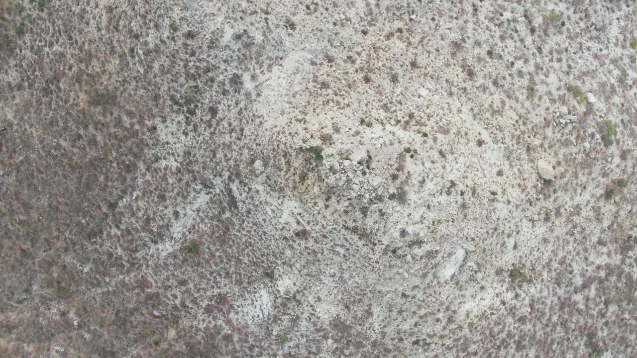 Aerial frame over Punta Gorda Beach showing mottled white soil, dark brown brush, a few green shrubs, and no visible people.