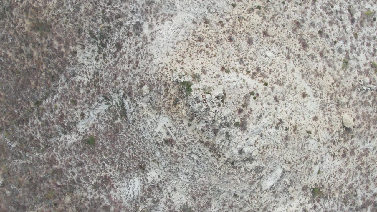 Aerial view of Punta Gorda Beach arid ground where pale soil bands mix with brown scrub, small shrubs, scattered stones, and no visible people.