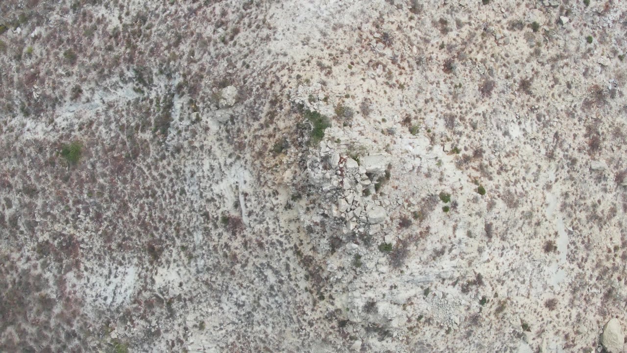Mid-clip overhead frame at Punta Gorda Beach, Baja California Sur, with gray-brown scrub, irregular stones, pale earth, and no visible people.