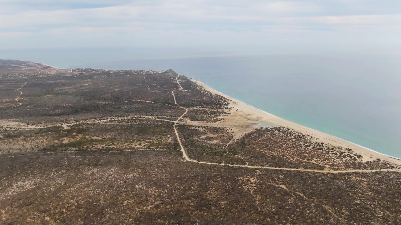 Aerial view of dirt tracks crossing dry coastal scrub beside a sandy beach and ocean.