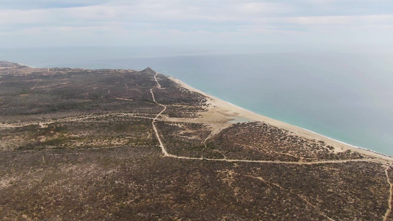 Wide aerial frame at Punta Gorda Beach with dry scrubland filling the lower frame, dirt paths crossing toward the sand, ocean to the right, and no visible people.