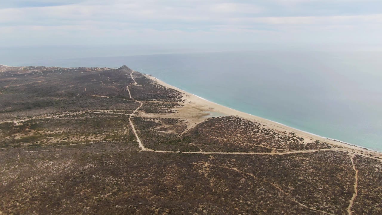 Aerial frame over Punta Gorda Beach, Baja California Sur, showing dry scrub terrain, pale dirt paths, a sandy shoreline, blue-green ocean, cloudy sky, and no visible people.